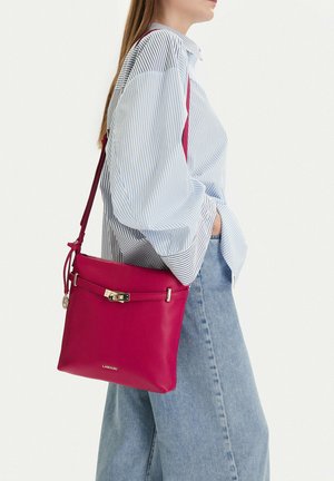 Frau trägt ein blau gestreiftes Hemd und hellblaue Jeans und eine leuchtend pinke Leder-Umhängetasche mit goldfarbenem Verschluss und Logo.