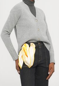 Cardigan gris en maille avec col en V et boutons, associé à un jean foncé. Une écharpe en soie blanche et jaune à motif est nouée à la taille.