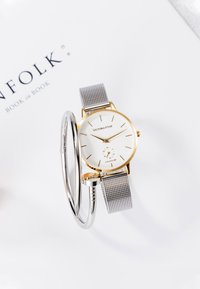 Harmaa verkko hihnalla varustettu kello, jossa on valkoinen kellotaulu ja kultasävyinen kotelo, jossa on kultaiset tuntimerkit ja pieni alikellotaulu. Esitetty renkaan kanssa.