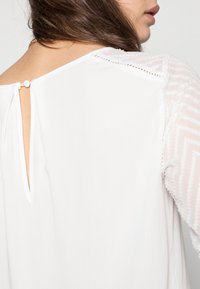 Blouse blanche avec une ouverture en forme de clé à l'arrière, fermeture à boutons et manches transparentes texturées avec des motifs en zigzag. Tissu doux.