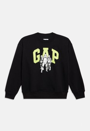 Svart genser med store gule "GAP"-bokstaver og tre hvite Star Wars Stormtrooper-figurer som går fremover under teksten.