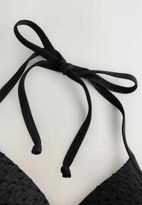 Haut de bikini noir texturé avec fines bretelles nouées en un nœud sur un fond clair.