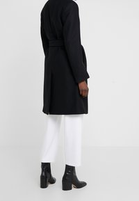 Cappotto nero con vita legata, maniche lunghe e taglio a metà lunghezza. Indossato sopra pantaloni bianchi e abbinato a stivali neri con tacco alto.