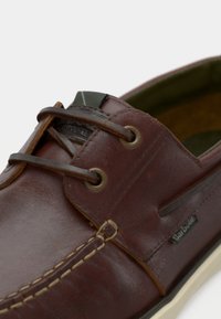 Καφέ δερμάτινο παπούτσι τύπου boat με ραφές, ορειχάλκινα κρικάκια και αντίθετη λευκή σόλα. Διαθέτει πράσινη υφασμάτινη λεπτομέρεια κοντά στα κορδόνια.