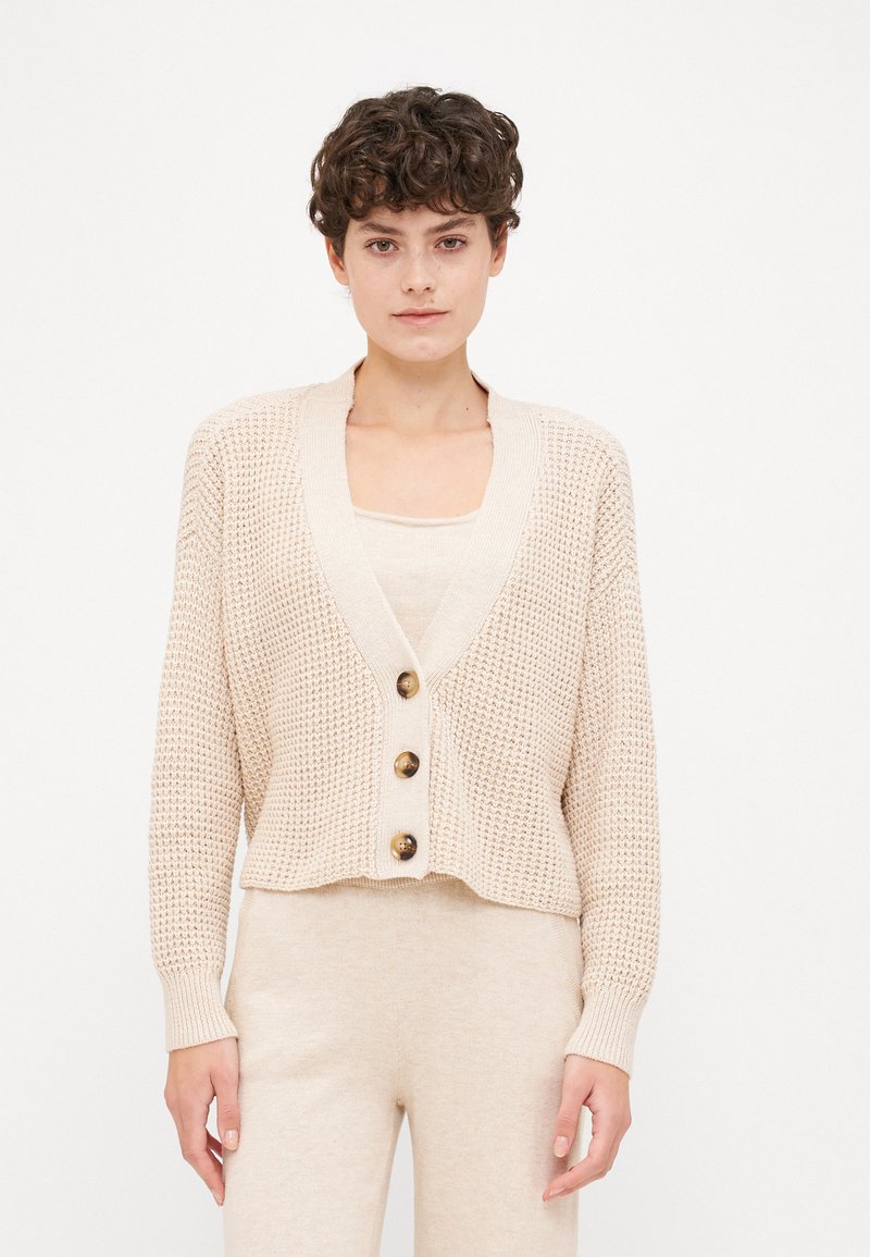 Beige knit cardigan with a V-neck, long sleeves, and four large buttons. Loose fit with a cropped hem and textured fabric.