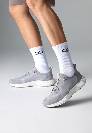 Person trägt weiße Socken mit schwarzen Unendlichkeitssymbolen und graue Sportschuhe mit strukturierten weißen Sohlen auf neutralem Hintergrund.