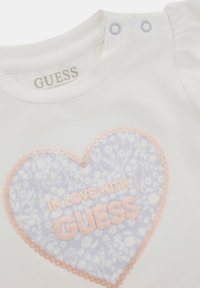 T-shirt bianco in cotone con un design a cuore floreale viola, bordato di paillettes e con la scritta "IN LOVE WITH GUESS" in caratteri rosa.