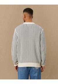 Hvit strikkegenser med vertikale svarte striper, ribbet krage og mansjetter, løs passform, kombinert med lyseblå denimjeans.