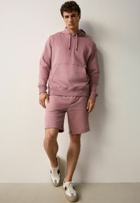 Sudadera rosa claro con un bolsillo frontal, combinada con pantalones cortos a juego. La tela es suave, con puños acanalados y capucha con cordón. Zapatillas blancas.