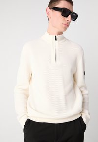 White ribbed sweater with a half-zip collar, featuring a textured pattern, complemented by black sunglasses and black pants.