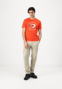Orange bomulds T-shirt med hvid og blå tekstgrafik, parret med beige cargo bukser og sorte støvler. Enkel, afslappet påklædning.