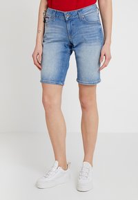 En person som bär en röd skjorta och blå denimshorts står i vita sneakers och poserar framför en enkel vit bakgrund.