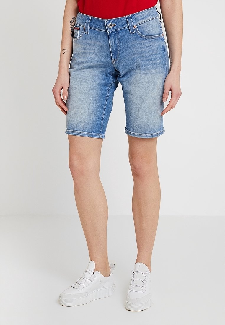 En person som bär en röd skjorta och blå denimshorts står i vita sneakers och poserar framför en enkel vit bakgrund.