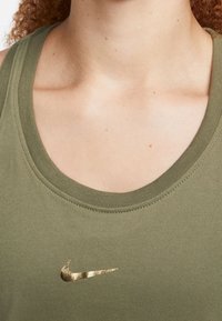 Oliwkowy top na ramiączkach z okrągłym dekoltem. Posiada złote logo Nike na piersi oraz teksturowane przeszycia wzdłuż szwów.