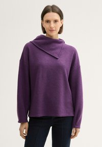 Lila, übergroßer Pullover mit hohem, gefaltetem Kragen, langen Ärmeln und strukturiertem Stoff. Kombiniert mit dunklen Jeans vor einem hellen Hintergrund.