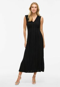 Abito maxi senza maniche nero con pieghe verticali, scollatura a V, tessuto fluido e una sottile texture. Indossato con tacchi bianchi con cinturini.