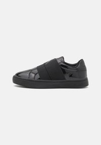 Black sneaker featuring a glossy patent finish with a wide elastic strap across the midfoot and a thick rubber sole. Minimalistic design.