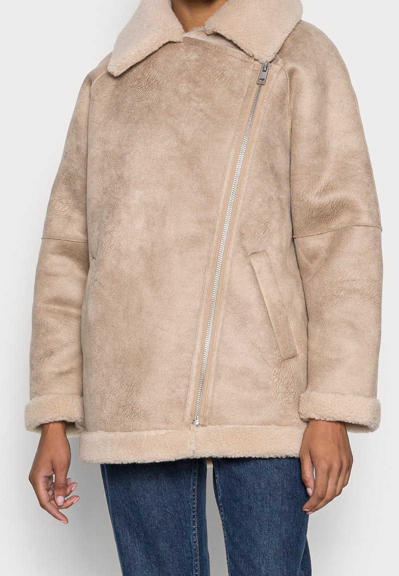 Personne portant une veste en shearling beige avec un grand col, une fermeture éclair argentée asymétrique, des manches remontées et un jean bleu.