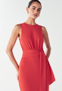 Rotes ärmelloses Kleid in figurbetontem Schnitt, mit gerafftem Oberteil und seitlichem Bindeband in der Taille. Glatter, strukturierter Stoff.