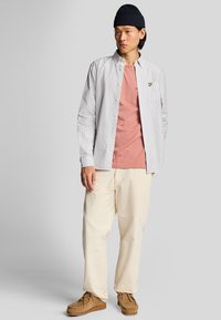 Camisa de botones a rayas claras sobre una camiseta rosa, combinada con pantalones crema y zapatos mocasín marrones. La persona lleva un gorro azul marino.