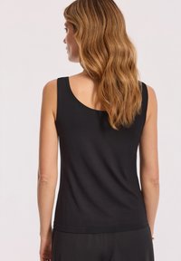 Kobieta z długimi falującymi, jasnobrązowymi włosami w czarnej, bezrękawnikowej bluzce i czarnych spodniach, pokazana od tyłu na jednolitym tle.