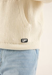 Cremfarbener gerippter Kapuzenpullover mit einer Tasche, mit einem schwarzen Logopatch am Saum, kombiniert mit hellblauen Jeans.