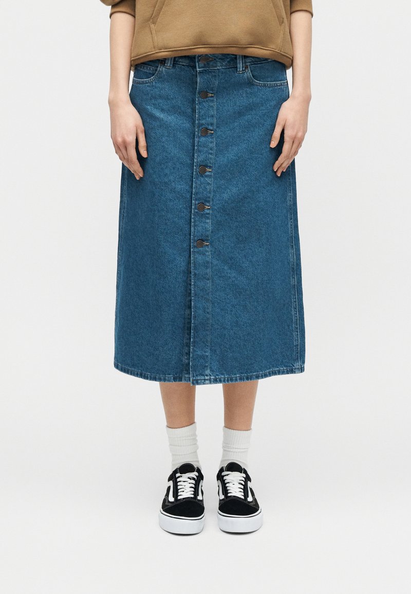 Frau trägt einen blauen Denim-Midirock mit Knopfleiste, beige Oberteil, weiße Socken und schwarze Turnschuhe mit weißen Schnürsenkeln, steht vor einem einfachen Hintergrund.