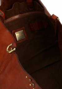 Interno di una borsa a tracolla in pelle marrone con una tasca con zip, un'etichetta dorata e rivestimento in tessuto scuro a contrasto; le caratteristiche includono dettagli in metallo e dettagli impresso.