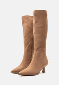 Un paio di stivali alti fino al ginocchio in suede beige con punte affusolate, cerniere laterali e tacco kitten basso su uno sfondo bianco.