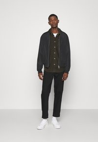 Zwarte bomberjack over een olijfgroene button-up shirt, gecombineerd met zwarte broek en witte sneakers. Gladde stof met een ritssluiting.