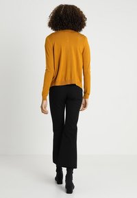 Person med lockigt hår som bär en senapsgul långärmad topp, svarta utsvängda byxor och svarta klackstövlar, stående mot en vit bakgrund.