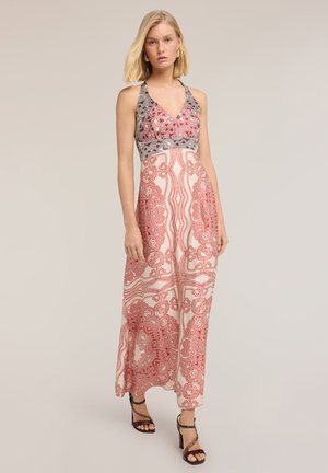 Vrouw staand, gekleed in een mouwloze rood-wit patroon halternek maxi-jurk met zwarte hakken sandalen tegen een effen achtergrond.