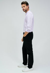 Pantaloni neri con vita regolabile, abbinati a una maglietta a maniche lunghe di un leggero colore viola. Il modello è in piedi di lato, indossando sneakers bianche.