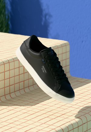 Basket en cuir noir avec semelle blanche, bout rond et accents texturés. Présente des lacets plats noirs et un marquage minimal sur le côté.