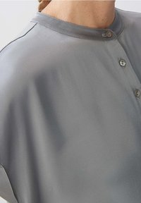 Gros plan d'une personne portant une chemise en soie grise avec un col mao et trois boutons le long de l'encolure avant.