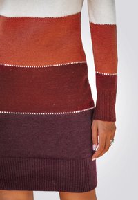 Gebreide trui met horizontale strepen in wit, oranje, rood en donkerpaars. Heeft ribbelboorden en een ribbelzoom, met decoratieve witte stiksels.