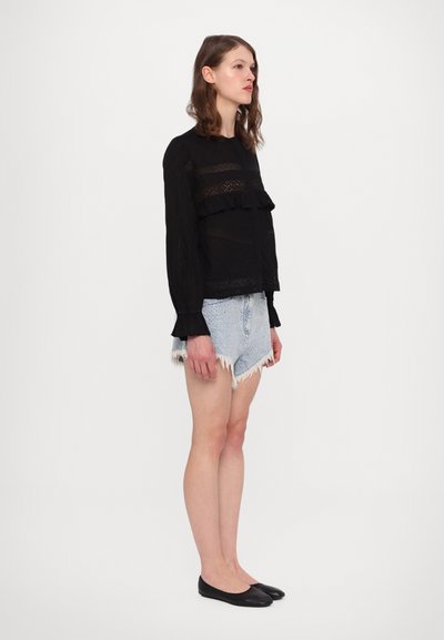 Schwarze Bluse mit transparenten Spitzen-Details und Rüschenelementen, kombiniert mit hellen Jeans-Shorts und schwarzen Ballerinas. Flach, schmal geschnitten.
