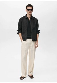 Black linen zip-up jacket, black knit top, cream trousers, and black sandals. Model wears dark sunglasses, standing against a plain background.