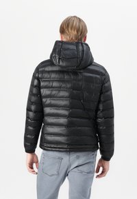 Zwarte pufferjack met een capuchon, voorzien van een gequilt ontwerp, glanzende afwerking en elastische manchetten. Draagt over lichtgrijze jeans, getoond vanuit de achterkant.