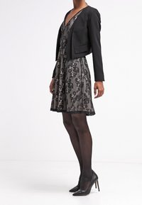 Veste noire courte portée sur une robe grise à motifs floraux, avec un décolleté en V et une jupe évasée. Portée avec des collants noirs transparents et des talons hauts.