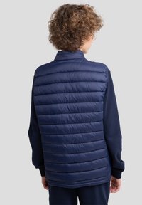 Gilet matelassé bleu marine avec un design matelassé. Il présente un col haut et un matériau léger, idéal pour superposer d'autres vêtements.