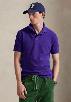 Mann trägt eine marineblaue Kappe, lila Polohemd mit grünem Logo und grüne Hose mit Kordelzug, steht vor neutralem Hintergrund.