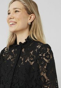 Blouse en dentelle noire avec des motifs floraux, col haut et manches courtes. Le modèle porte des boucles d'oreilles rondes en argent et a de longs cheveux ondulés.