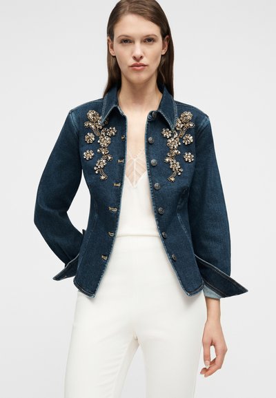 Γυναίκα που φοράει σκούρο τζιν μπουφάν με χρυσή κεντητή λουλούδια, πάνω σε λευκό τοπ με δαντέλα και ψηλόμεσο λευκό παντελόνι, στέκεται μπροστά σε απλό φόντο.