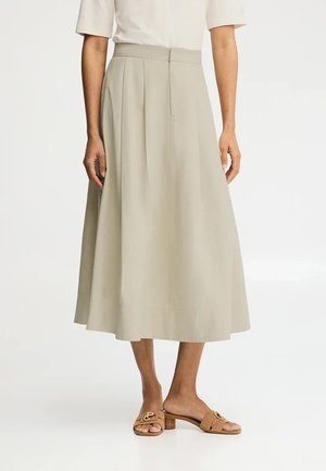 Mujer con falda midi beige con pliegues, combinada con sandalias abiertas de tacón bajo en color marrón claro, de pie sobre un fondo blanco.