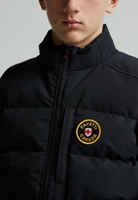 Giubbotto imbottito nero con colletto alto, presenta una chiusura con zip e una patch rotonda gialla con la scritta "ZAVETTI CANADA" e un design di foglia d'acero.
