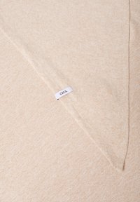 Tissu tricoté beige avec une texture subtile et un motif chiné. Présente une forme triangulaire et une étiquette "CECIIL" visible.