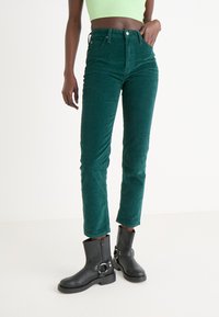 Zelene kord traperice viskog struka sa slim fit krojem, s prednjim dugmetom i zatvaračem, uparene s crnim čizmama do gležnja s kopčama.