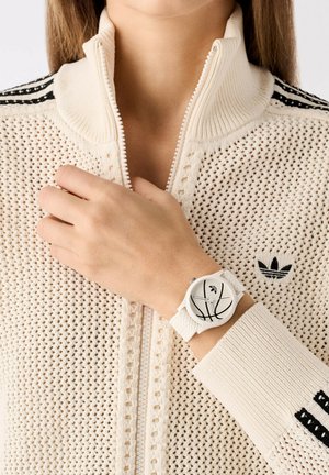 Montre de sport blanche avec un cadran rond présentant un motif de basketball, portée sur une main au-dessus d'un pull zippé en crème avec un logo.