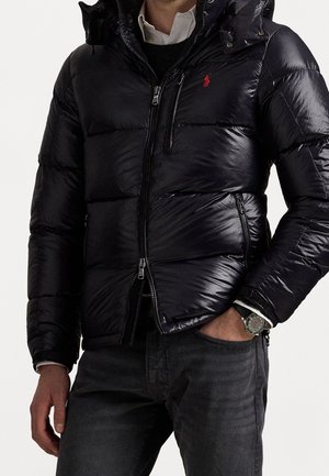 Homme portant une doudoune noire brillante avec des poches zippées, une chemise blanche, un pull noir, un jean foncé et une montre-bracelet.